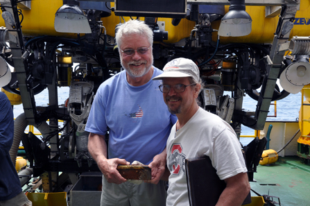 Recovery Limited Partnership's director of operations Craig Mullen (left) and chief scientist & historian Bob Evans (right) hold one of the five gold bars recovered by Odyssey's Remotely Operated Vehicle ZEUS (background) during the first reconnaissance dive to the SS Central America shipwreck in April 2014. Copyright: Odyssey Marine Exploration, Inc., www.OdysseyMarine.com.