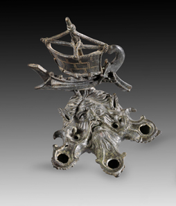 Lot 234: Oceanus-shaped bronze lamp with attachment in the shape of a ship. Roman, 1st-2nd cent. AD. Lamp in the shape of bearded Oceanus’ head with crab claws. At the head attachment in the shape of a Roman galley. From the Shlomo Moussaieff Collection, since 1948, Israel. With export permission from the Israel Antiquities Authority. Black-green patina, mast and edge of sail broken, mast repaired with a pin, tip of mast missing, parts of ropes missing, small hole on one nozzle. Estimate: 50,000,- euros. Hammer price: 55,000 euros.