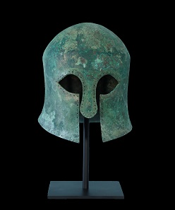 An ancient Greek bronze helmet of the Corinthian type, with curvilinear eye holes that taper to a point, a wide nose guard, broad cheek pieces that leave a vertical opening for the mouth, and a border that has been drilled with holes to secure an interior lining. Traces of the original crest are evident in the patina.